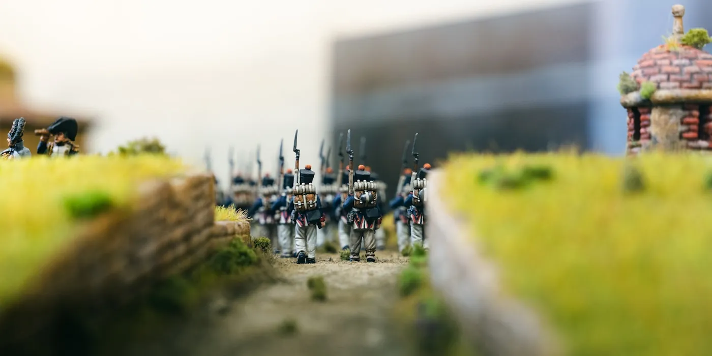 A regiment of historical infantry miniatures marching through a tabletop diorama between stone walls.